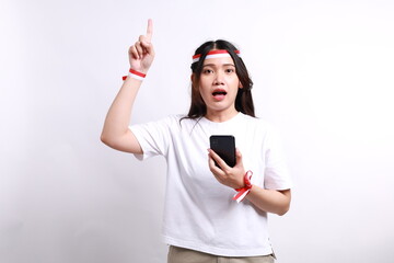 Surprised young Asian woman with red white flag ribbon holding mobile phone and pointing a finger up isolated over white background. Celebrate Indonesian independence day on 17 August