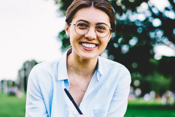 Portrait of cheerful young woman in spectacles enjoying learning outdoors sitting on grass in park,...