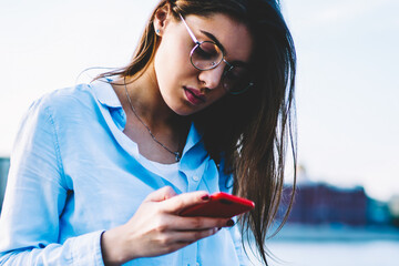 Pensive hipster girl in eyewear reading news from web page on smartphone resting with coffee to go...