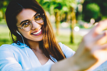 Portrait of cute cheerful girl in eyewear making selfie on modern smartphone camera enjoying...