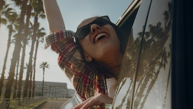 Woman in sunglasses with happy smile waves hand out of car window riding along road with palm trees. Cheerful long-haired girl expresses pure delight enjoying summer trip on hot summer day