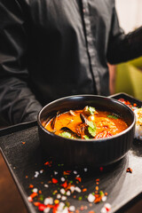 Close-up of a bowl of spicy seafood soup garnished with herbs, presented by a person in a dark attire, against a blurred background
