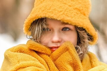 A blonde teenage girl in a yellow jacket and hat walks through the spring city. Wrapped in a coat against the cold