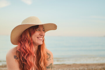 A woman with red hair and a straw hat is smiling at the camera. She is sitting on the beach, looking out at the ocean