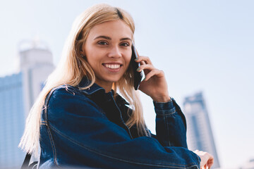 Portrait of carefree hipster girl with blonde hair smiling at camera while having mobile...