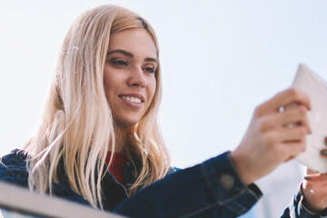 Smiling female tourist in denim wear making photos on modern touch pad device to share in social networks via 4G internet.Blonde young woman taking picture for own blog during trip standing on street