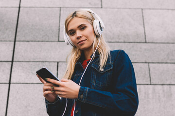 Below portrait of blonde attractive female blogger listening audio songs in modern headphones while...
