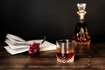 There is a decanter and a glass of cognac on a wooden table, and an open book with a flower lies on...