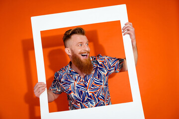 Photo of impressed funky guy with red beard wear print shirt hold frame look at offer empty space isolated on orange color background