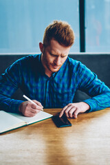 Pensive smart student rewriting information from website using internet connection on smartphone sitting in coworking.Young man with red hair noting records from telephone in notepad studying remotely