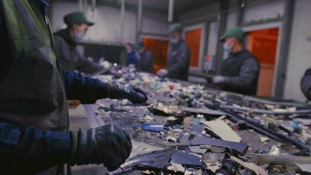 Male workers of a trash recycling plant sort through garbage on a rubbish conveyor belt. Men take waste with their hands or palms. Recyclables are moving in line at the garbage factory.