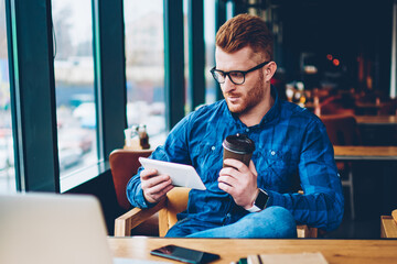 Stylish male blogger with red hair reading websites with last news on modern tablet connected to free 4G internet resting with coffee to go in cafeteria interior sitting at table with laptop computer