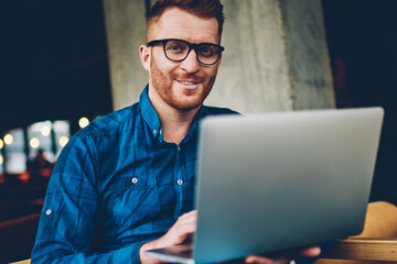 positive bearded male graphic designer with red hair and eyeglasses smiling at camera while working...