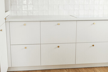 Modern kitchen interior. Stylish white kitchen cabinets with brass knobs, granite countertop, and...