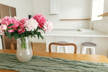 Beautiful peonies in vase on wooden table on background of stylish white kitchen with appliances in new scandinavian house. Modern kitchen interior and summer floral arrangement