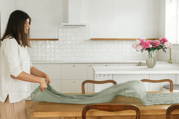 Woman arranging green textile table runner on wooden table in new modern home. Young female...