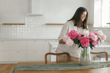 Woman holding beautiful peonies in vase on wooden table in new modern home. Young female decorating...
