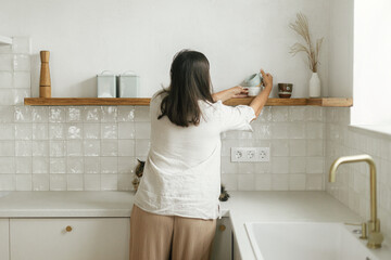 Stylish woman unpacking and decorating shelves with utensils in new minimal white kitchen. Housewife cleaning up kitchen after moving in in new modern scandinavian home