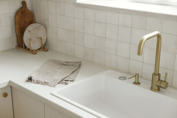 Stylish white kitchen cabinets, brass faucet and granite sink, wooden shelves with utensils  in new scandinavian house. Modern minimal kitchen design. Modern kitchen interior