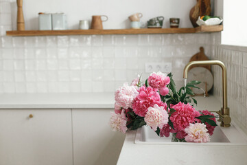 Pink peony flowers in modern kitchen interior, summer floral arrangement. Beautiful peonies in sink on background of brass faucet and white counter in new scandinavian house