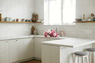 Beautiful peonies in sink on background of brass faucet and window in new kitchen in scandinavian house. Pink peony and roses flowers in modern kitchen interior, summer floral arrangement