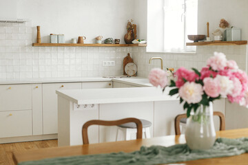 Modern kitchen interior. Beautiful peonies in vase on wooden table and stylish white kitchen with appliances in new scandinavian house. Minimal kitchen design and summer floral arrangement