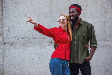 Young cheerful couple, where a man and a blonde woman stand embracing on a background of a gray wall and looking away where the girl points