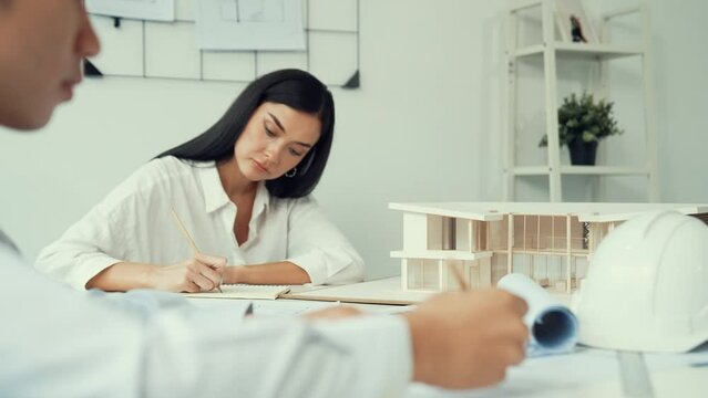 Professional architect engineer drawing blueprint and working together with young beautiful caucasian coworker on meeting table with safety helmet, house model, blueprint scatter around. Immaculate.