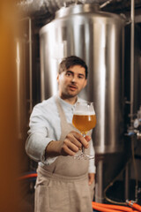Happy brewer. Happy young male brewer in apron holding glass with beer and looking at it with smile while standing in front of metal containers
