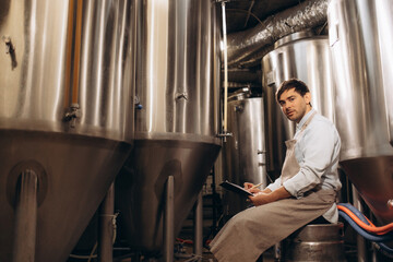 Portrait of brewery worker checking fermentation process, technology and app for small business. Concentrated millennial attractive man worker in apron, with tablet controls boiler in plant interior