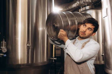 Young man in leather apron holding beer keg at modern brewery, craft brewery worker