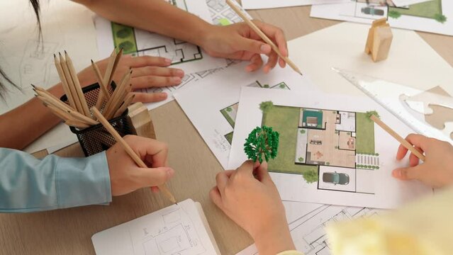 Architect and engineer use tree model discuss about building blueprint on table with architectural document, measurement tools and wooden block scatter around at modern office. Closeup. Delineation.