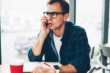 Young good looking emotional male student in cool eyeglasses having mobile conversation via...