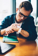 Young handsome pondering skilled freelancer in cool eyewear planning working schedule using digital smartwatch sitting indoors.Pensive hipster guy checking updates on modern device in coffee shop