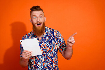 Portrait of impressed man with red long beard hold tablet indicating at offer empty space isolated on vibrant orange color background