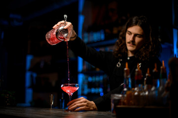 Bartender pours a cocktail from a mixing glass into a stemmed glass