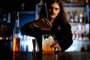 Bartender with long hair stirs a cocktail with ice in a glass with a bar spoon