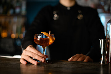 Bartender holds a glass with a cocktail by the stem, to which a piece of zest is attached with a...