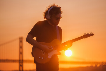 Hipster street rock musician in black playing electric guitar in street outdoors on sunset