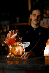 Focus on cocktail glass standing in flames on bar counter with bartender pouring cocktail from mixing glass
