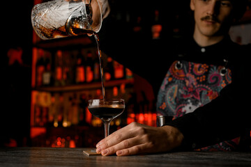 Bartender pours a brown cocktail from a mixing glass with an ice strainer inserted