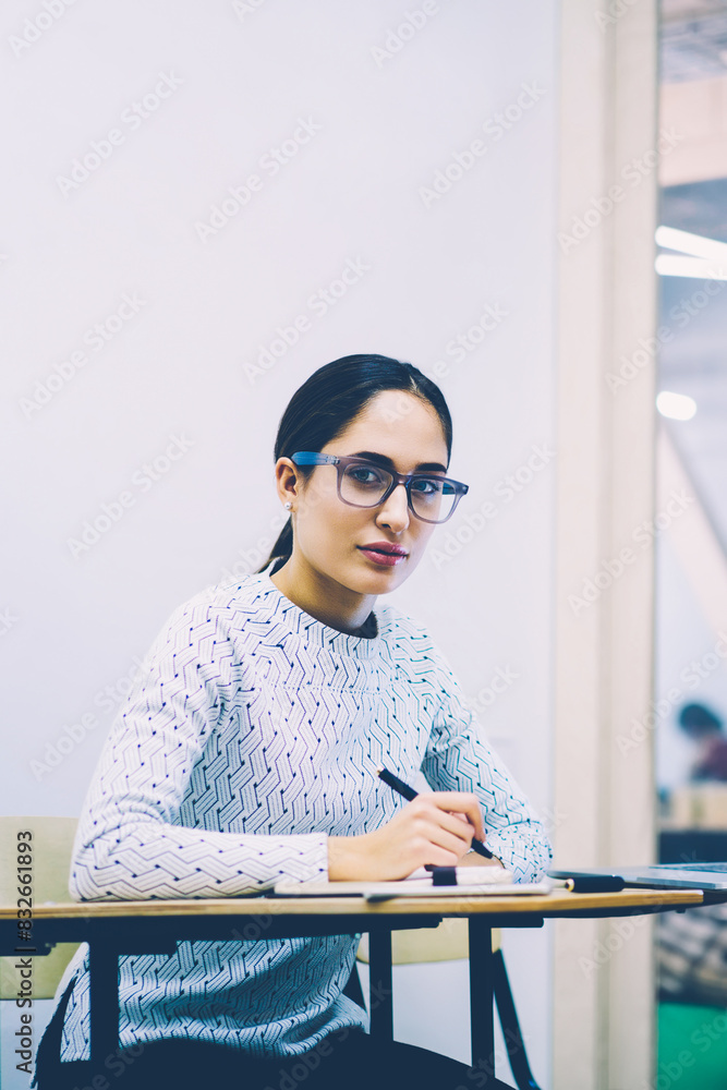 Poster portrait of serious female student in eyewear doing homework task writing report in classroom,concen