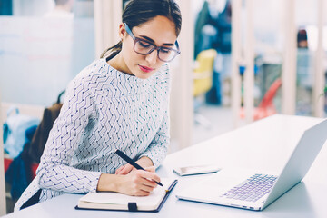 Pensive woman in eyewear planning working schedule writing in notebook while sitting at working...