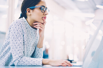 Side view of pensive female administrative manager in eyewear standing on working place organizing...