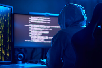 In a dimly lit room, a cloaked figure types code on a computer, hinting at a mysterious hacker