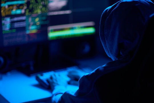 In a dimly lit room, a cloaked figure types code on a computer, hinting at a mysterious hacker