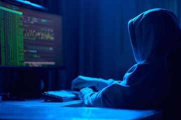 In a dimly lit room, a cloaked figure types code on a computer, hinting at a mysterious hacker