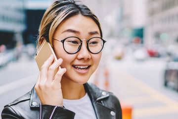 Young asian female in eyewear having mobile conversation while walking in big city center, crossing...