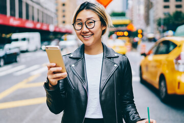 Positive asian woman walking with coffee to go using application for calling taxi and paying online on smartphone, smiling chinese girl reading information from web page on mobile passing avenue