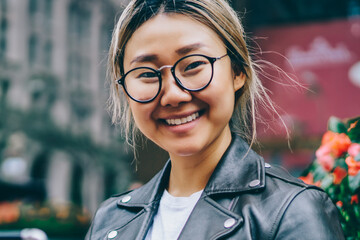 Portrait of cheerful asian female in eyewear resting on bench on square blogging in social networks...